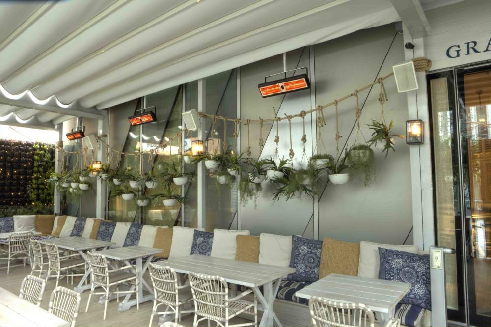 Infrazářič Tansun Monaco Single 1,5 kW v instalaci na restaurační terase – ukázka prémiového Low Glare quartzového zářiče s minimální svítivostí a zlatým reflektorem ve večerní atmosféře s tlumeným osvětlením, kde zářič nenarušuje estetiku prostoru
