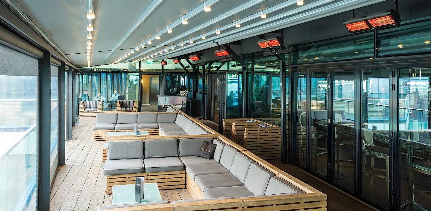 Infrazářič Tansun Monaco Single 1,5 kW v instalaci na restaurační terase – ukázka prémiového Low Glare quartzového zářiče s minimální svítivostí a zlatým reflektorem ve večerní atmosféře s tlumeným osvětlením, kde zářič nenarušuje estetiku prostoru