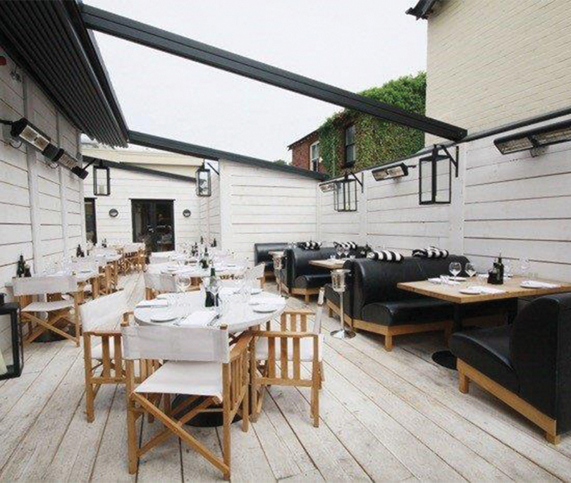 Infrazářič Tansun Monaco Single 1,5 kW v instalaci na restaurační terase – ukázka prémiového Low Glare quartzového zářiče s minimální svítivostí a zlatým reflektorem ve večerní atmosféře s tlumeným osvětlením, kde zářič nenarušuje estetiku prostoru